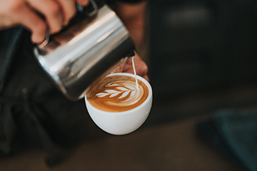 Milch wird aus einem Edelstahl-Milchkännchen in einen Cappuccino gegossen